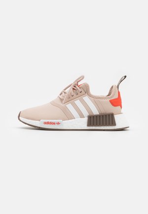 Baskets de sport beige clair avec des rayures blanches, des accents orangés et une semelle extérieure en caoutchouc texturé. Dotées d'un col rembourré et d'un système de laçage.