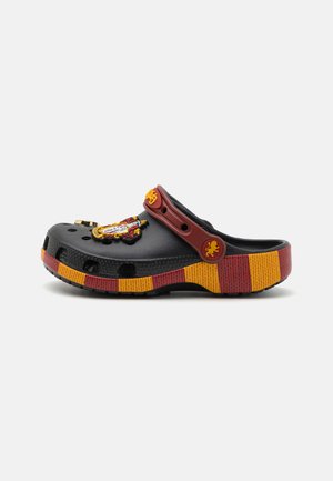 Schwarze Clogs mit einer gelb-roten gestreiften Sohle, featuring einem Löwenemblem und verstellbarem Riemen. Hergestellt aus leichtem synthetischem Material.