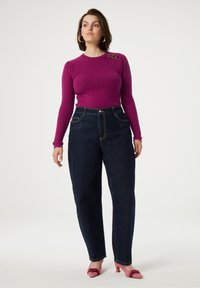 Fuchsia ribbelgebreide trui met lange mouwen en drie goudkleurige accenten, gecombineerd met donkerblauwe high-waisted jeans met geborduurde details.