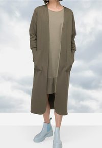 Cardigan verde alargado con frente abierto y dos bolsillos, usado sobre un vestido verde claro, combinado con botines azules hasta el tobillo.