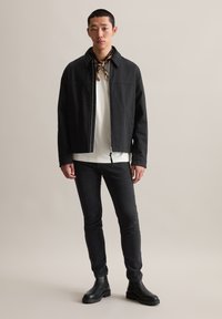 Schwarze Jacke mit Kragen, Reißverschluss und zwei seitlichen Taschen, kombiniert mit einem weißen T-Shirt, schwarzen Jeans und schwarzen Knöchelstiefeln.