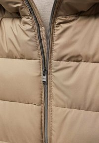 Gros plan d'une veste matelassée beige avec une fermeture éclair argentée partiellement fermée et un tissu beige en dessous.