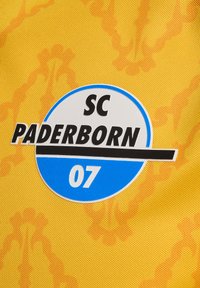 Λογότυπο της SC Paderborn 07 σε μαύρο, μπλε και άσπρο χρώμα σε υφασμάτινη επιφάνεια κίτρινου χρώματος με διακριτά πορτοκαλί σχέδια.