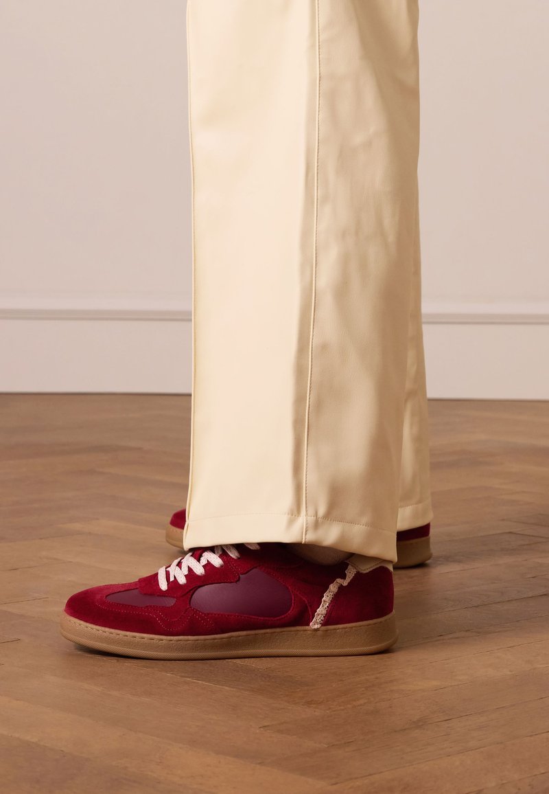 Rode suède sneakers met een gladde rubberen zool, voorzien van een wijnrode accent, witte veters en decoratieve stiksels. Gekaapt met crèmekleurige broek.