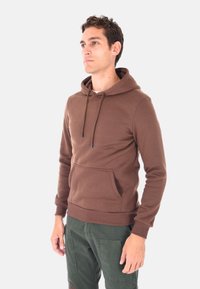 Uomo che indossa un felpa con cappuccio marrone con tasca frontale e pantaloni cargo verde scuro, in piedi e che guarda di lato su uno sfondo bianco.