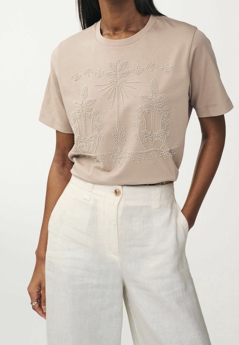 Femme portant un t-shirt beige à manches courtes avec des motifs floraux en perles de nacre, rentré dans un pantalon en lin blanc taille haute.