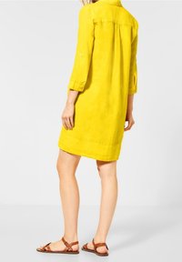 Vestido de lino amarillo con cuello, mangas tres cuartos y diseño de solapa en la parte posterior, combinado con sandalias marrones de tiras. Textura suave.