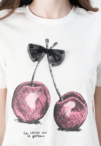 Hvid T-shirt med en håndtegnet illustration af to pink kirsebær med sorte stilke, accentueret af en sort, gennemsigtig sløjfe med polka dots.