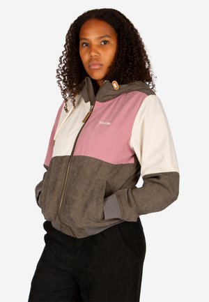 Frau mit lockigem Haar, die eine Cordjacke mit Reißverschluss und braunen, pinken und cremefarbenen Farbblöcken trägt und die Hände in den Taschen hat.