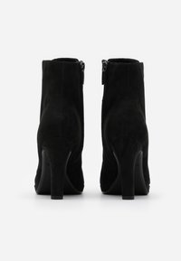 Botas de tobillo de ante negro con tacón alto, que cuentan con cremalleras laterales y una textura suave y mate. La forma general es estilizada y ajustada.