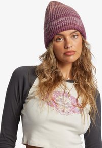 Gorro en degradado púrpura, hecho de tela de punto acanalado. Presenta una parte superior cónica y un pequeño etiquetado con el logotipo. El modelo lleva una parte superior en crema y gris.