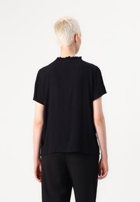 Schwarzes Kurzarmoberteil aus strukturiertem Stoff mit gerüschtem Ausschnitt und lockerem Schnitt, kombiniert mit schwarzen Hosen für einen monochromen Stil.