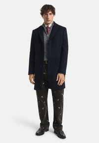 Uomo che indossa un cappotto blu navy, un maglione grigio con zip, una cravatta rossa a motivo, pantaloni in corduroy scuri con stampe di uccelli e scarpe di pelle nere su uno sfondo bianco.