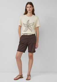 Lichtgele katoenen t-shirt met een zeesterafbeelding, gecombineerd met bruine katoenen shorts en een crèmekleurige geweven riem. Bruine sandalen met gekruiste banden.