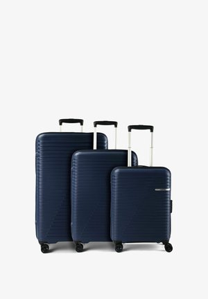 Trois valises rigides bleu marine de tailles différentes avec des poignées télescopiques déployées et quatre roulettes pivotantes chacune, disposées côte à côte sur fond blanc.