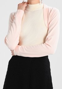Top de cuello alto de color crema con textura acanalada y mangas largas de color rosa suave; tejido ligero con una textura suave, encima de una falda negra con textura.