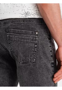 Jean gris foncé avec un design structuré. Comprend une poche arrière et des coutures visibles. Le matériau semble texturé, avec des détails subtils.