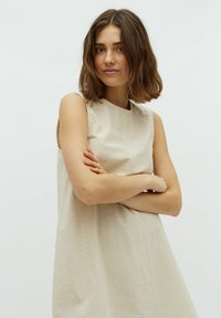 Junge Frau mit schulterlangem braunem Haar, die ein ärmelloses beige Kleid trägt und mit verschränkten Armen vor einem schlichten weißen Hintergrund steht.