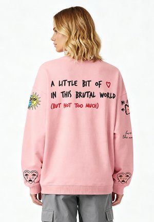 Sweatshirt surdimensionné rose clair avec du texte et des graphiques. Présente un design de soleil sur une manche et une illustration de cœur avec des larmes.