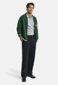 Mann steht mit den Händen in den Taschen, trägt eine grüne Reißverschlussjacke über einem grauen Polohemd, marineblaue Hose und schwarze Schuhe.