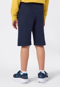Enfant portant un short bleu marine, des chaussettes blanches Champion et des baskets bleu marine avec des accents bleus, se tenant de dos face au spectateur.