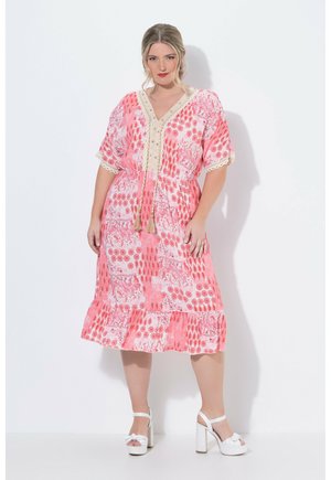 Kvinde i en pink mønstret midi-kjole med blonder trimmet V-hals, kvastbånd og hvide højhælede platformsandaler står mod en hvid baggrund.