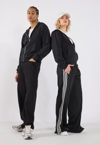 Svart hettegensere med kengurulommer, kombinert med svarte bukser; en har hvite sidestriper. Begge har på seg joggesko og caps.