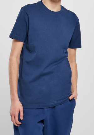 Homme portant un t-shirt bleu marine uni à manches courtes et un pantalon assorti, une main dans la poche et l'autre le long du corps.