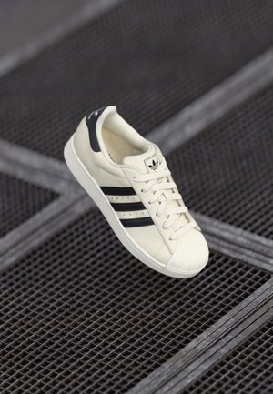 Biały sneaker Adidas z czarnymi paskami na tle metalowej siatki.