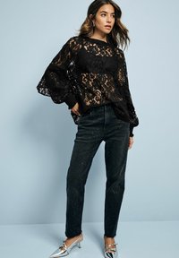 Černý krajkový top se širokými rukávy, kombinovaný s černými džíny s tečkovaným vzorem. Modelka má na sobě stříbrné lodičky na podpatku. Pozadí je světle modré.