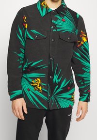 Czarna kurtka lekka z polarowej tkaniny z zielonym wzorem liści palmowych, czerwonymi akcentami i żółtymi detalami. Posiada dwie kieszenie na piersi oraz zapięcie na guziki.