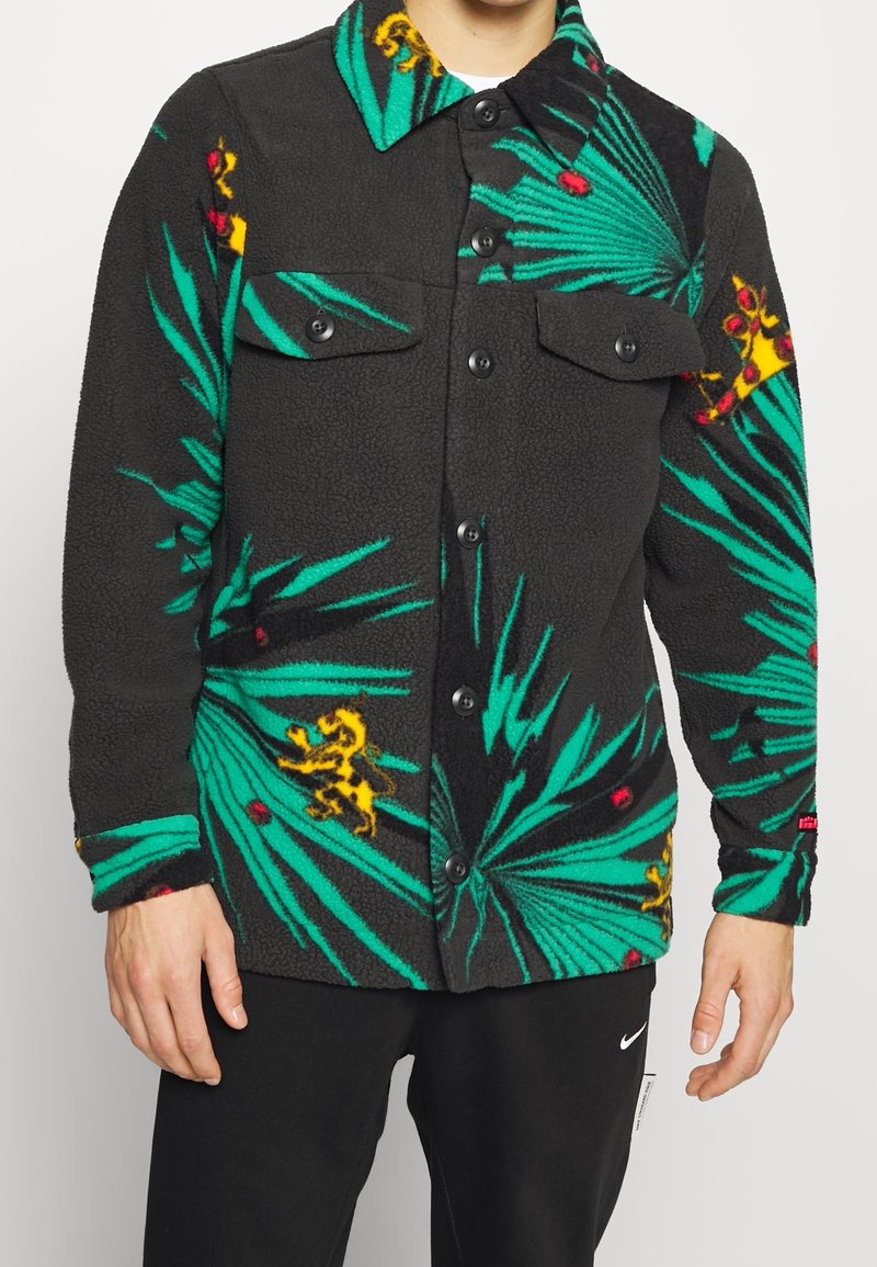 Czarna kurtka lekka z polarowej tkaniny z zielonym wzorem liści palmowych, czerwonymi akcentami i żółtymi detalami. Posiada dwie kieszenie na piersi oraz zapięcie na guziki.