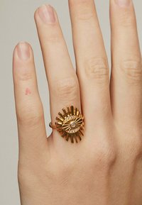 Anillo de oro con un diseño de estallido solar y un motivo de ojo, adornado con una piedra central y piedras de acento a lo largo de los bordes, exhibido en una mano.