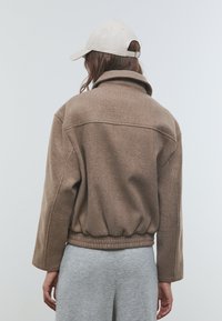 Veste en mélange de laine marron avec un col montant, une taille élastique et des épaules tombantes. Associée à un pantalon gris à taille élastique. Fond clair.