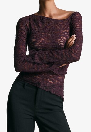 Persona que lleva una blusa de manga larga de encaje translúcido en púrpura oscuro con los brazos cruzados, combinada con pantalones negros de cintura alta.