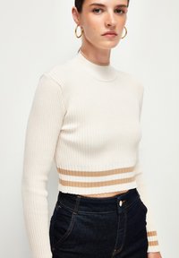 Kremowy prążkowany top w stylu crop z wysokim dekoltem i długimi rękawami. Posiada poziomą paskowaną lamówkę w kolorze beżowym i białym. Połączony z ciemnymi dżinsami.