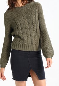 Pull en maille vert olive avec un motif de câble texturé, associé à une jupe en denim noir avec une fente sur le devant et un ourlet effiloché.
