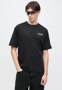 Μαύρο κοντομάνικο t-shirt από λείο ύφασμα με λαιμόκοψη τύπου crew και μικρό λευκό γραφικό λογότυπο στην αριστερή πλευρά. Φορεμένο με σκούρο τζιν.