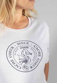 Witte t-shirt met een ronde blauwe afbeelding van een zeepaardje en de tekst: "CACHE COEUR SINCE 2008 OCEAN ATLANTIQUE" op de voorkant.