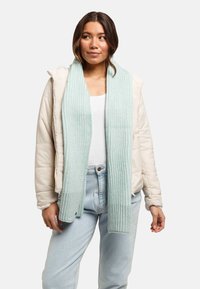Veste matelassée beige clair avec capuche, accompagnée d'une écharpe douce en maille côtelée vert menthe. L'ensemble comprend un haut blanc et un jean bleu clair.