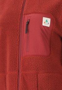 Rote Fleecejacke mit einem Reißverschlusstaschen auf der Brust, glatten Stoffakzenten und einem kleinen Logopatch auf der oberen linken Seite.