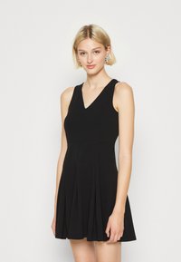 Une robe noire sans manches avec un décolleté en V, un corsage ajusté et une jupe plissée évasée, fabriquée à partir d'un tissu doux au toucher lisse.