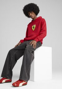 Rød hættetrøje med gult Ferrari-logo, grå vidde bukser og røde Puma-sko. Modellen har en afrofrisure og poserer på en hvid kube.