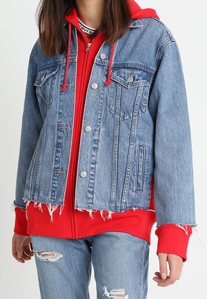 Giacca di jeans con orlo sfilacciato, colore blu, due tasche al petto, indossata sopra un hoodie rosso con coulisse, abbinata a jeans blu strappati.