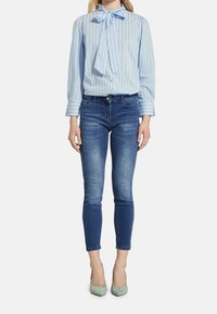 Kvinna i blårandig blus med slips, mörkblå tajta jeans och ljusgröna högklackade skor, som står mot en vit bakgrund.