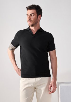Polo negro de manga corta con una tela texturizada, un cuello ventilado y un diseño ajustado, combinado con pantalones de color claro.