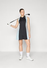 Golfové šaty s zipem, černým tělem a světle modrými stranami. Styling s límečkem a v kombinaci s bílými botami a golfovou rukavicí.
