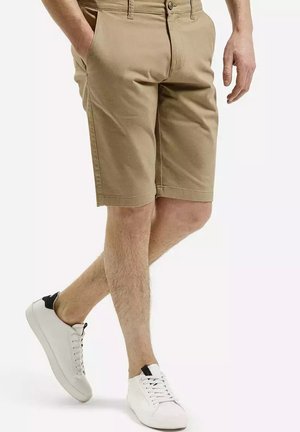 Khaki szorty z bocznymi kieszeniami, płaskim przodem i szlufkami na pasek, w połączeniu z białymi butami sportowymi na sznurówki z czarnym akcentem na pięcie.