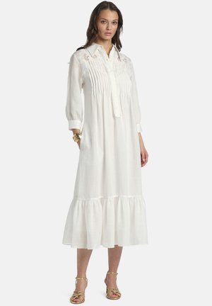 Kobieta ubrana w białą midi sukienkę z długim rękawem, ozdobioną koronkowymi detalami, z plisami z przodu, zapinaną na guziki, z falbanistym dołem oraz złotymi sandałami na paski.