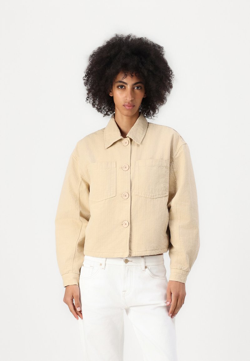 Jaqueta curta bege, com gola, grandes bolsos frontais e fecho de botão. Tecido suave e texturizado, com um corte descontraído.